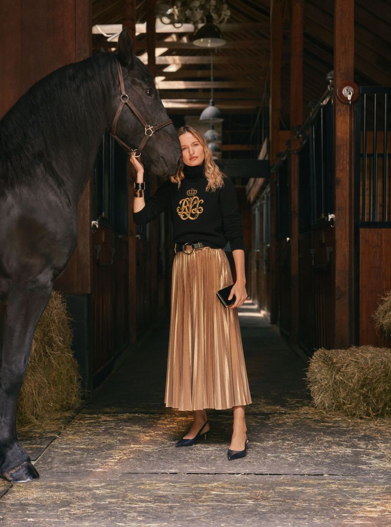 Woman in a black top and gold skirt poses with a black horse in a barn, creating a fashion editorial shot with warm tones.