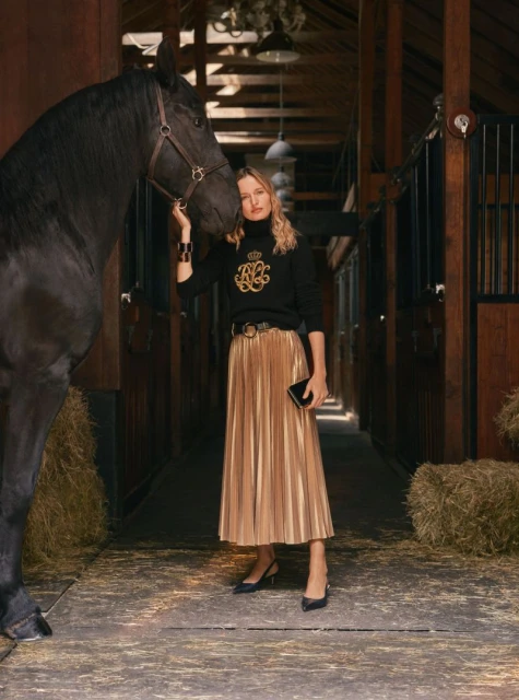Woman in a black top and gold skirt poses with a black horse in a barn, creating a fashion editorial shot with warm tones.