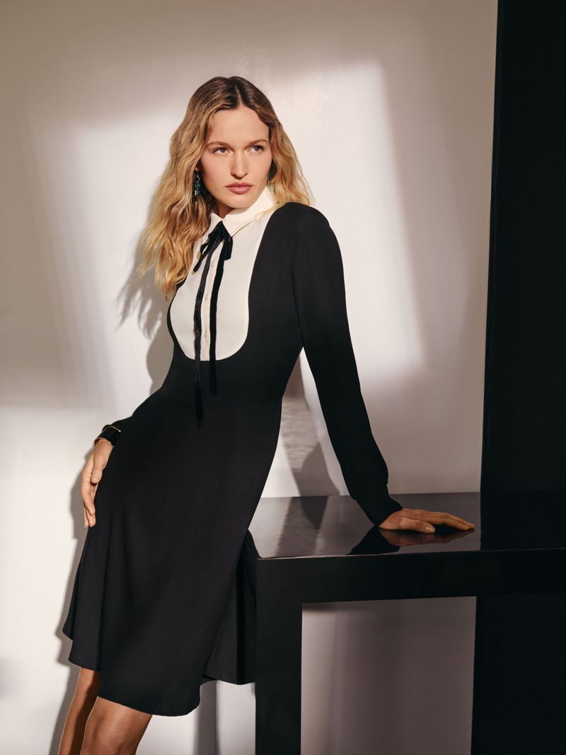 Elegant adult woman in a black long-sleeve dress with a white collar and bow, posing indoors against a neutral backdrop with a black table.