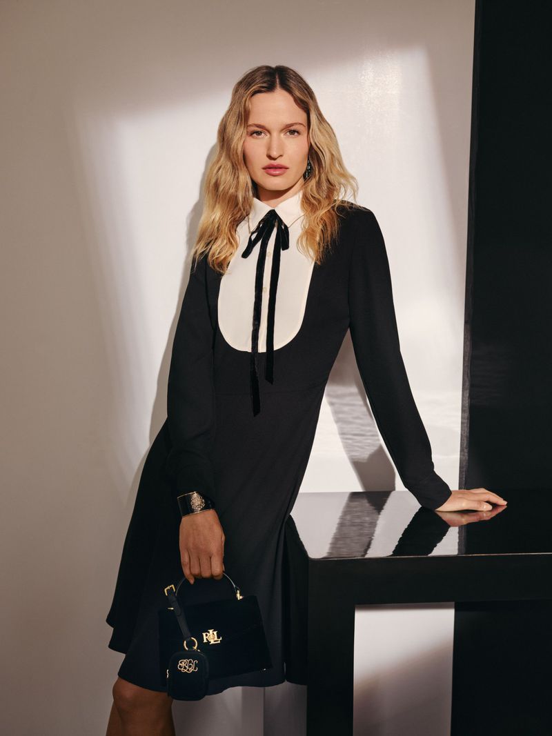 A stylish adult female model poses in a black and white dress with a black purse in a studio with contrasting light and dark areas.