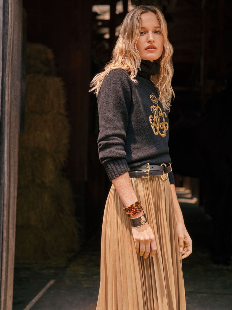 Woman poses in black turtleneck with gold embroidery, pleated skirt, and brown accessories against a rustic backdrop.