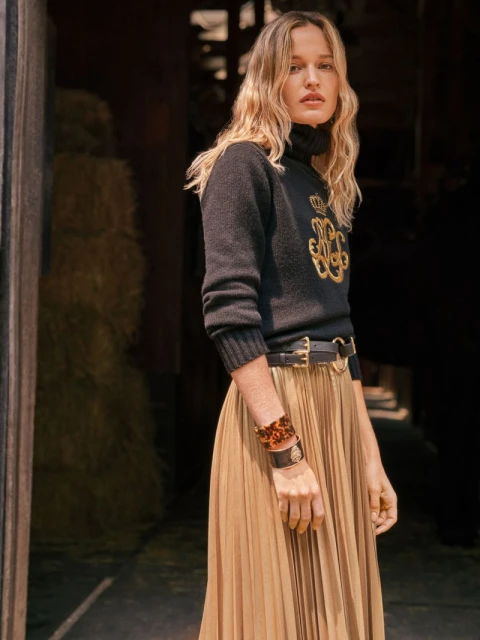 Woman poses in black turtleneck with gold embroidery, pleated skirt, and brown accessories against a rustic backdrop.