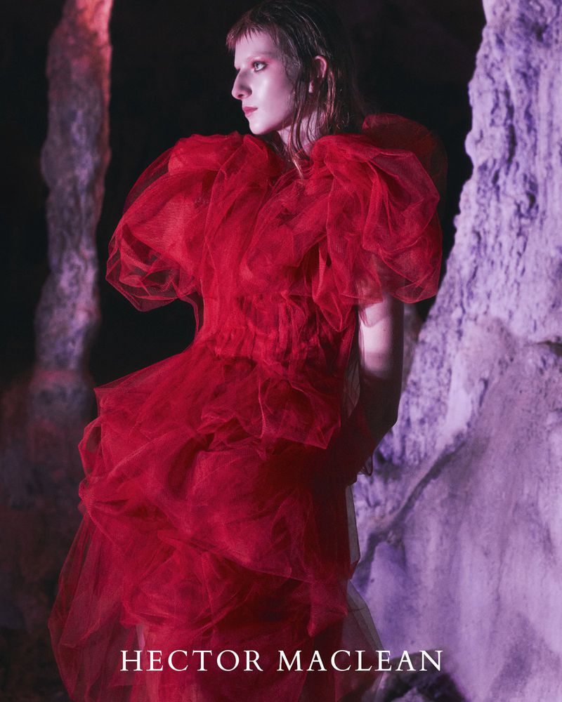 Elegant woman poses in a red tulle gown in front of a textured rock wall, illuminated by purple light and a dark background.