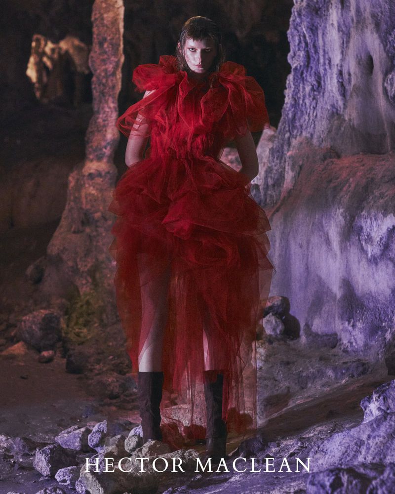 Adult female fashion model in a red tulle gown stands in a cave. The image is low-lit with soft colours and interesting rock formations.
