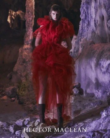 Adult female fashion model in a red tulle gown stands in a cave. The image is low-lit with soft colours and interesting rock formations.