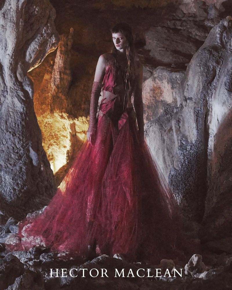 A fair-skinned woman with long brown hair poses in a red gown with a tulle skirt in a cave with rocks and dappled light.