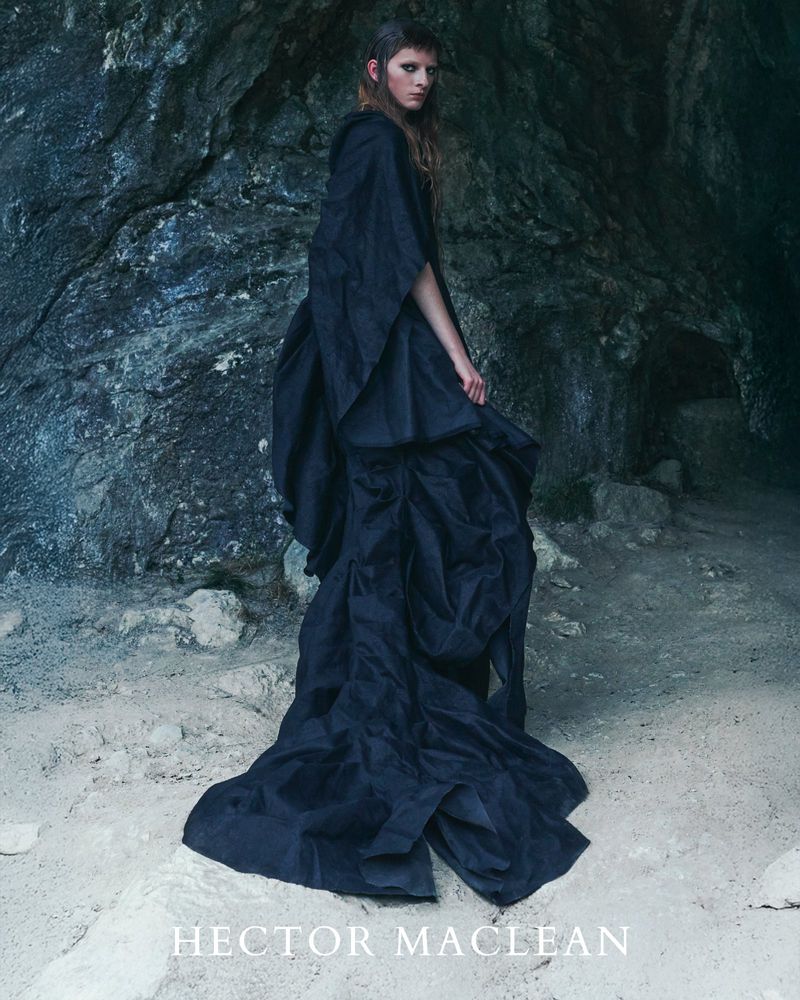 Fashion shot of a woman in a dark gown posing in a cave with a stone background and natural lighting, showcasing editorial style and design.