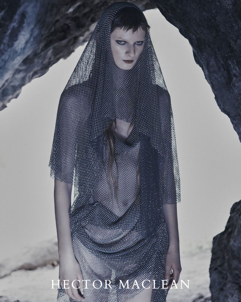 Fashion portrait of a woman wearing a sheer, silver mesh veil and dress, standing in front of a stone cave-like structure.
