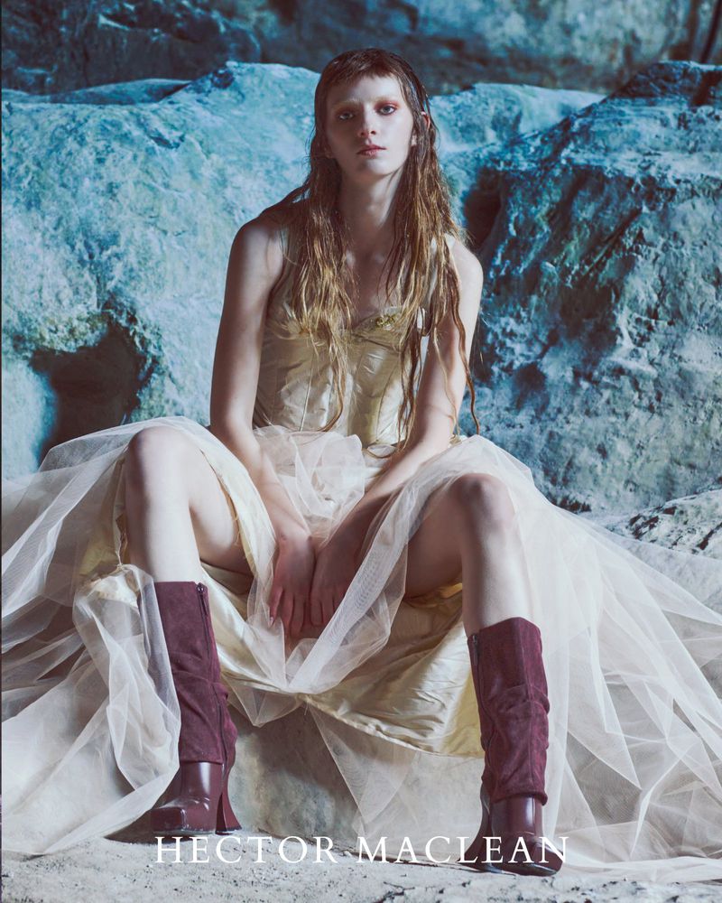 A fair-skinned model poses in a cream-colored dress and burgundy boots against a rock background, exuding editorial fashion.
