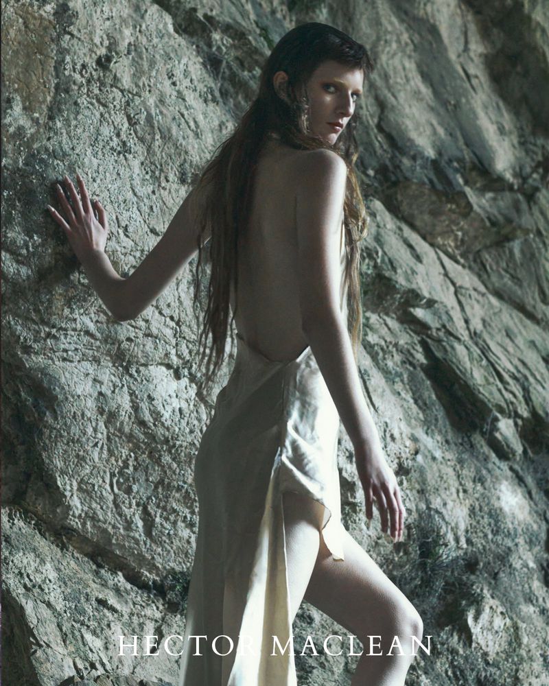 Woman in flowing cream gown poses against a rugged stone wall, her gaze directed at the viewer, creating a stark and evocative image.