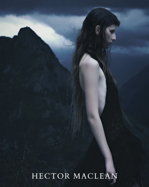 A solitary, fair-skinned woman with long dark hair wearing a black dress stands in a mountainous landscape with a cloudy sky above.