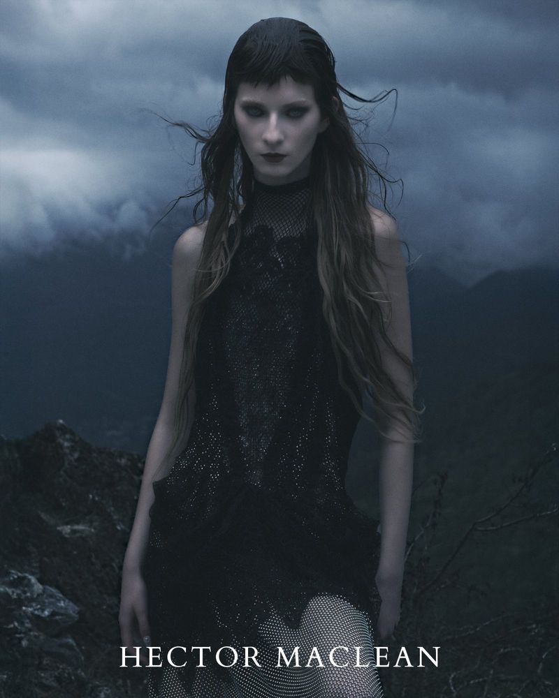 Fashion photo of a woman with long brown hair wearing a black dress, posing in front of a dark, cloudy sky and mountainous terrain.