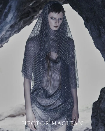 Model wearing a sheer silver chainmail hooded dress standing inside a rocky cave opening for Hector Maclean S/S 26