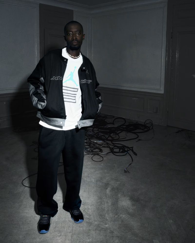 An adult male stands indoors, wearing a jacket with a logo, a white t-shirt, black pants, and black shoes. Cables are on the floor.