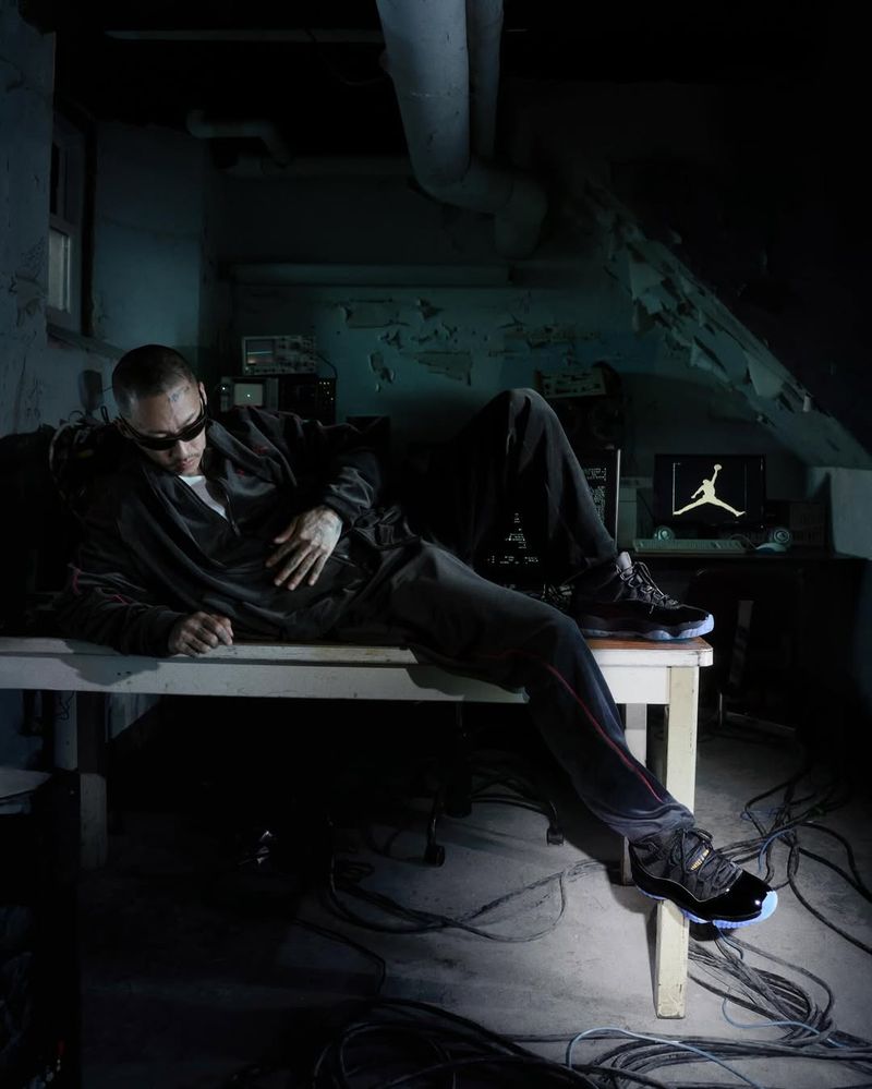 Man in sunglasses lounges on a white table in a dimly lit room, showcasing sneakers with a vintage computer and screen background.
