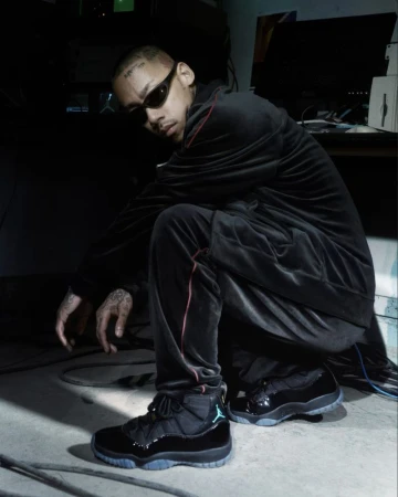 Man in a black tracksuit and sunglasses crouching indoors, wearing black sneakers, with computer equipment visible in the background.