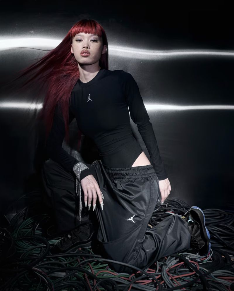 Woman with red hair kneeling in front of a metallic background wearing a black long-sleeve shirt and black athletic pants with sneakers.