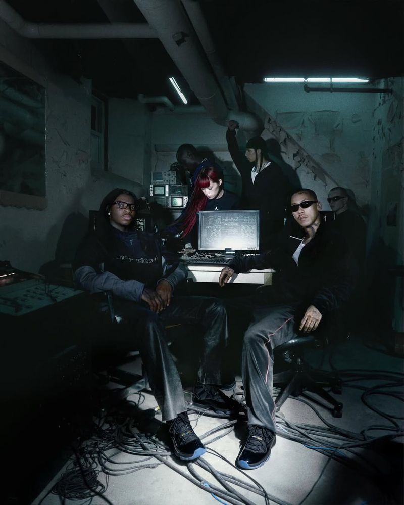 Group of seven people in a dark room with computers and wires. Two men sit in the foreground wearing dark clothing and sneakers.