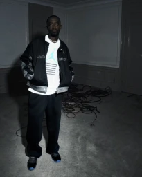 Man standing in dim room wearing black jacket, white graphic t-shirt, and dark sweatpants for Nike Jordan campaign.