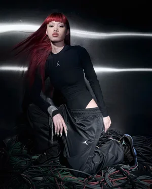 Model with bright red hair wearing a black long-sleeve top and trousers kneeling on tangled cables against a metallic background