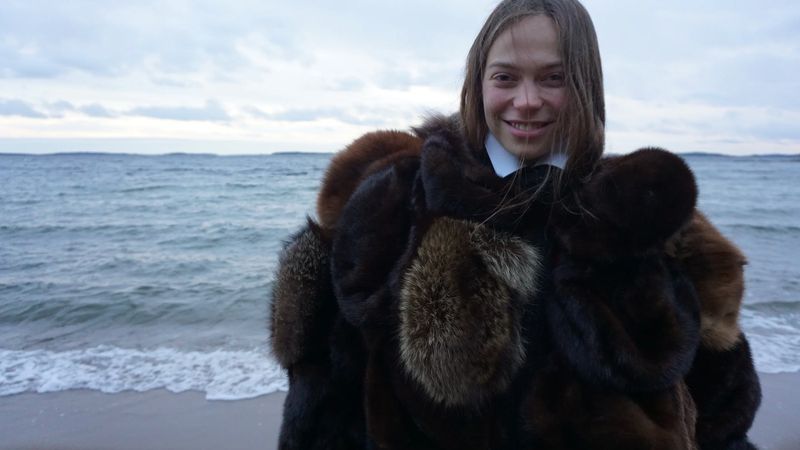 Smiling woman in a fur coat poses on a sandy beach with ocean and cloudy sky behind her on a cloudy day. Wide shot.