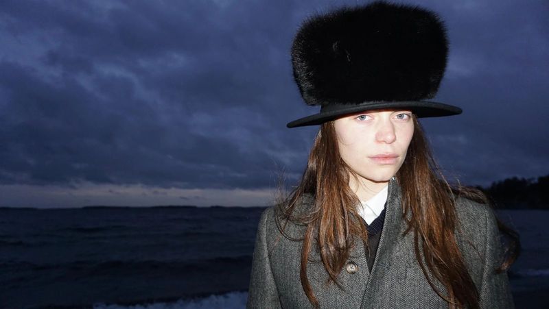 Portrait of a woman in a fur hat and gray coat standing on a beach with stormy sky, wearing neutral makeup, looking at the camera.