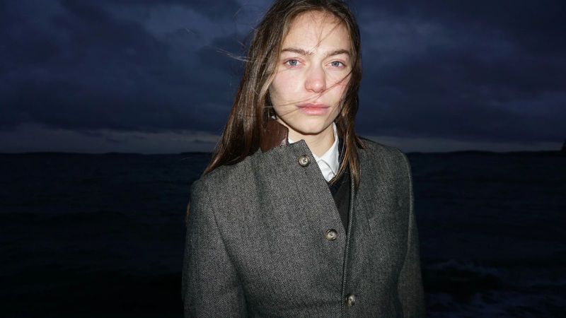 Portrait of a brunette woman in gray tweed jacket stands outdoors with blurred seascape background, wind blowing hair on a dark night.
