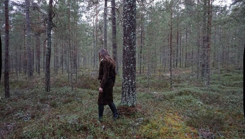 Adult female with long brown hair walks through a dense pine forest wearing a brown patterned jacket and black leggings in daylight.