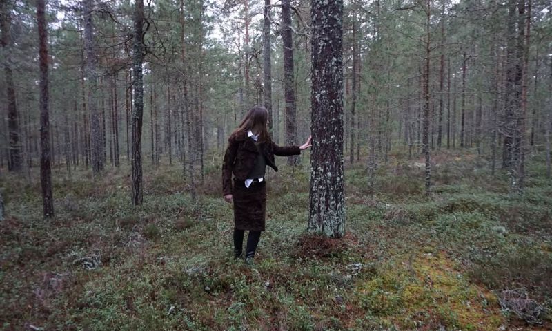 Woman in a brown suit stands in a pine forest touching a tree, surrounded by greenery in a natural setting with tall trees.