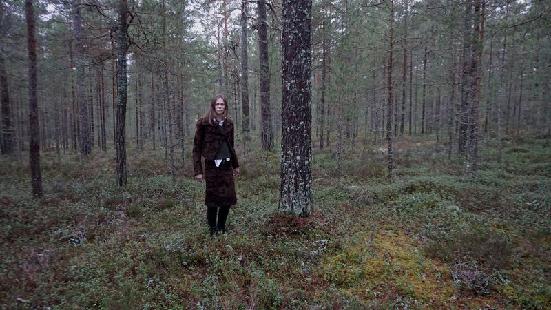 Woman in brown patterned skirt and jacket stands in a lush green forest with tall pine trees, under an overcast sky.