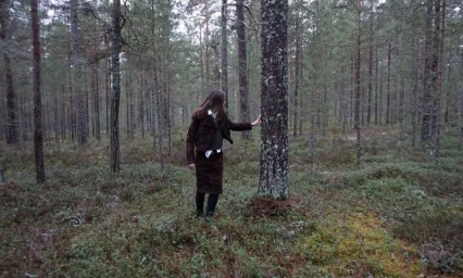A woman in a brown jacket and skirt standing in a dense pine forest, touching a tree trunk.