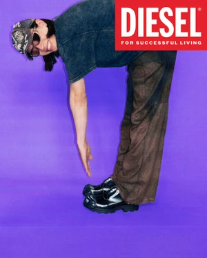 Model bending forward wearing dark distressed jeans, t-shirt, cap, sunglasses and shiny shoes against a bright purple backdrop