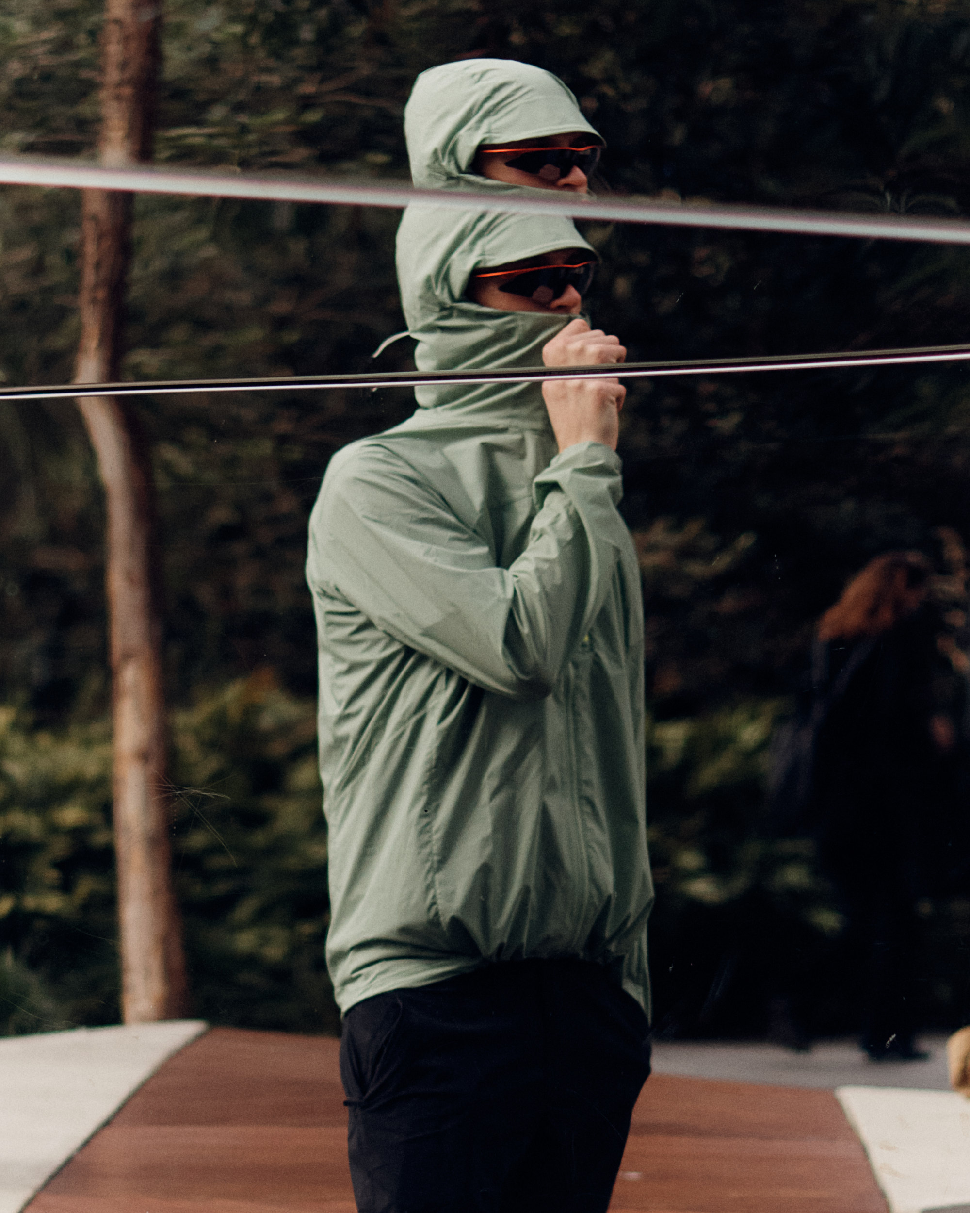 A figure in a hooded, lightweight jacket and sunglasses stands outside with trees in the background, partially reflected in a surface.