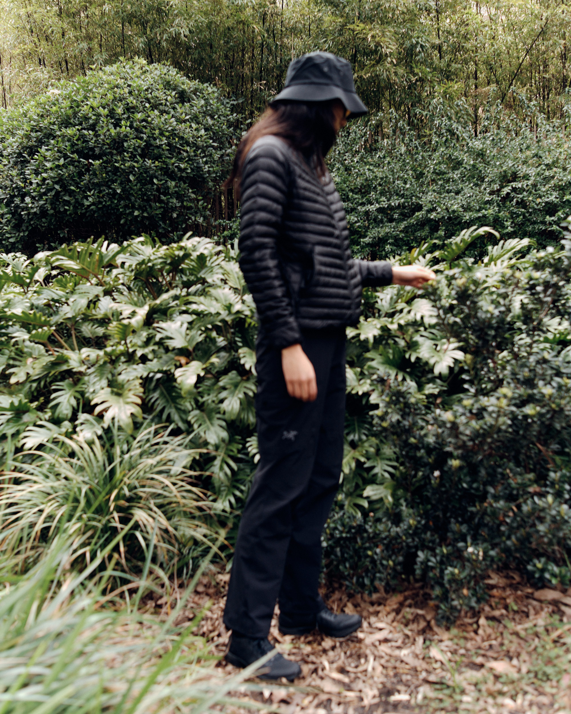 Woman in black puffer jacket and bucket hat stands amid lush greenery. Natural light illuminates the outdoor scene.
