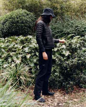 Woman in black puffer jacket and bucket hat stands amid lush greenery. Natural light illuminates the outdoor scene.