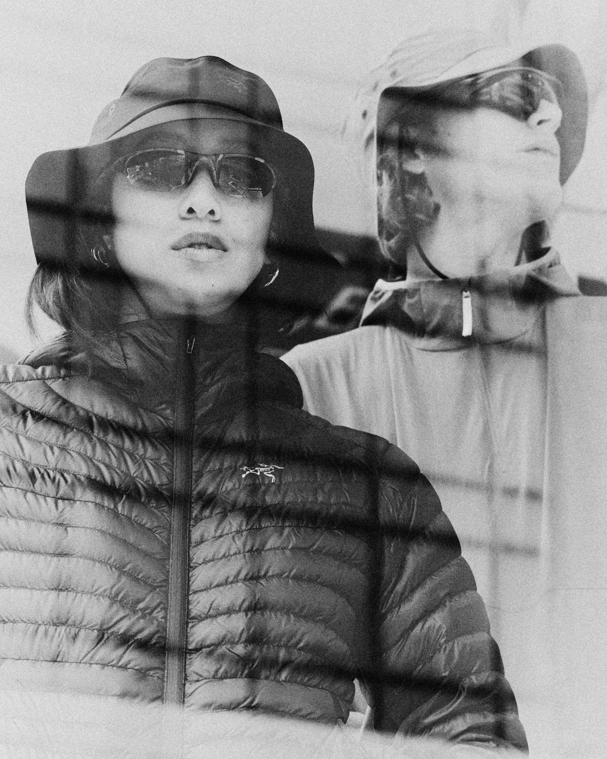 Black and white portrait of two models wearing puffer jackets and bucket hats, shot through a window grid.