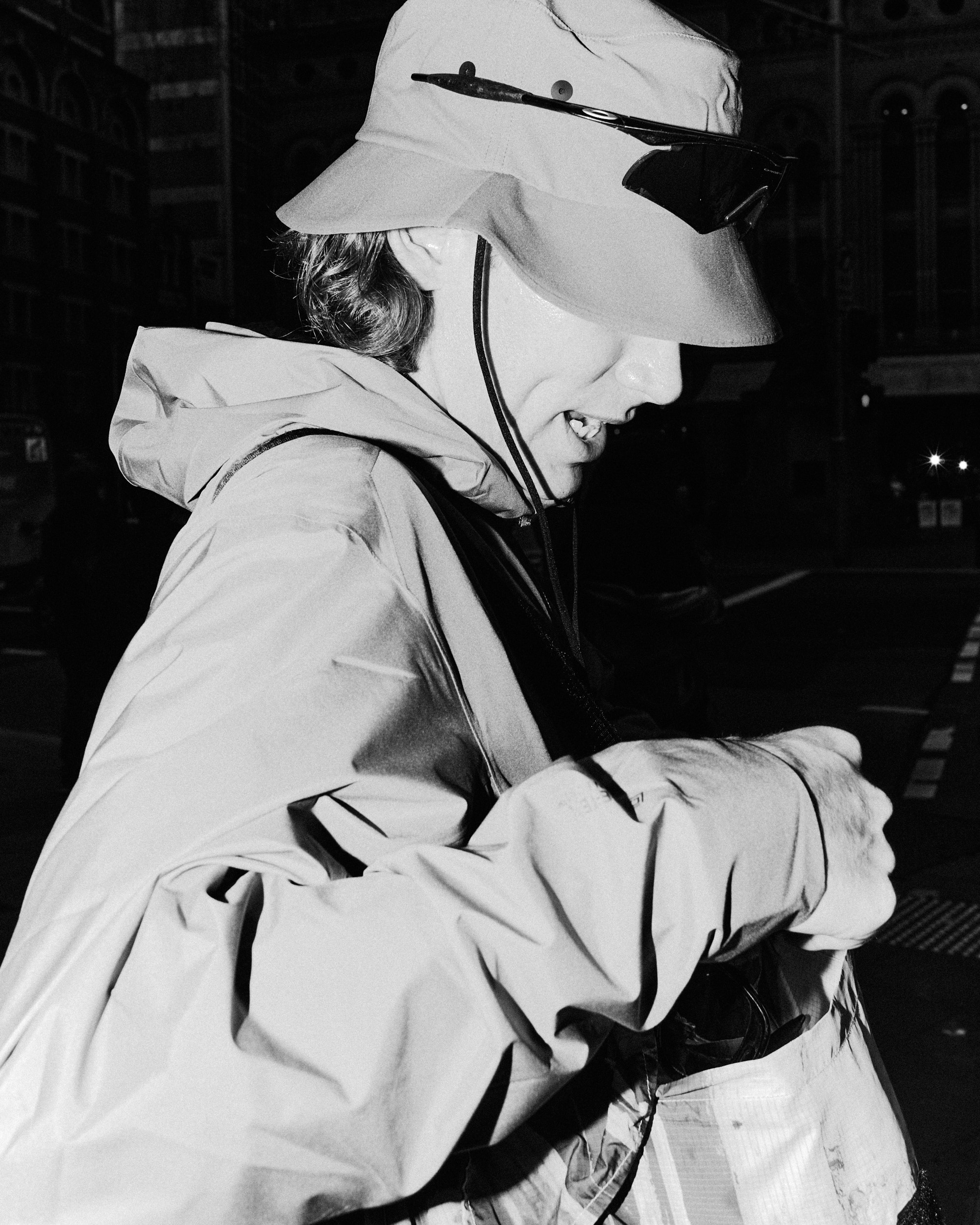 Black and white portrait of an adult wearing a hat and jacket, looking down and holding an object outdoors at night.