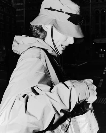 Black and white portrait of an adult wearing a hat and jacket, looking down and holding an object outdoors at night.