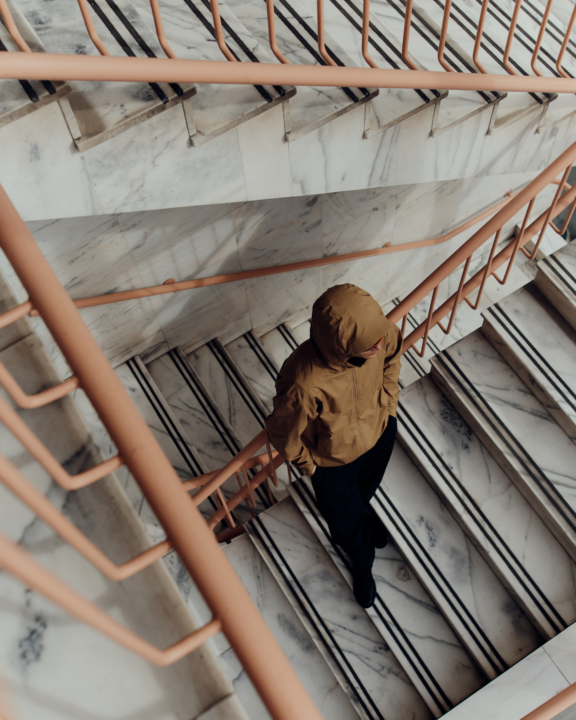 High-angle shot of a hooded person walking down stairs with a marble finish and peach-colored metal railing in an indoor setting.