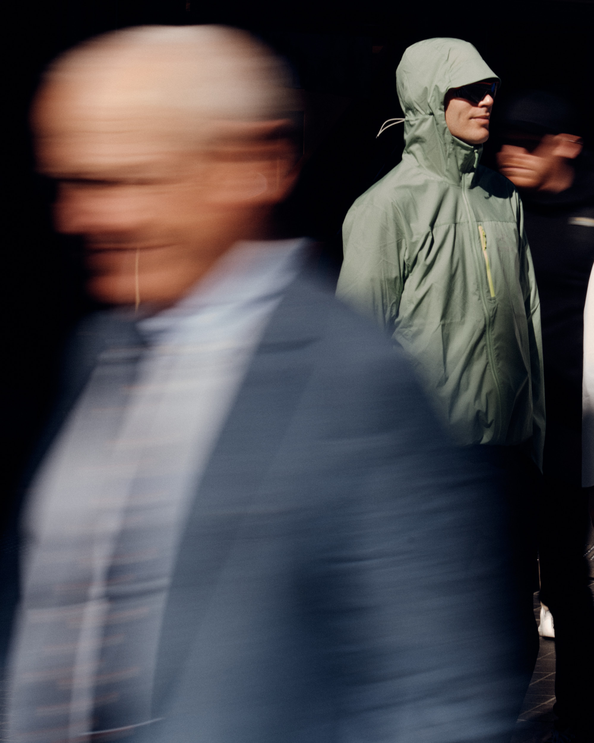 Three people walking, one out of focus, another in a light green jacket with a hood, captured in an outdoor setting with natural light.