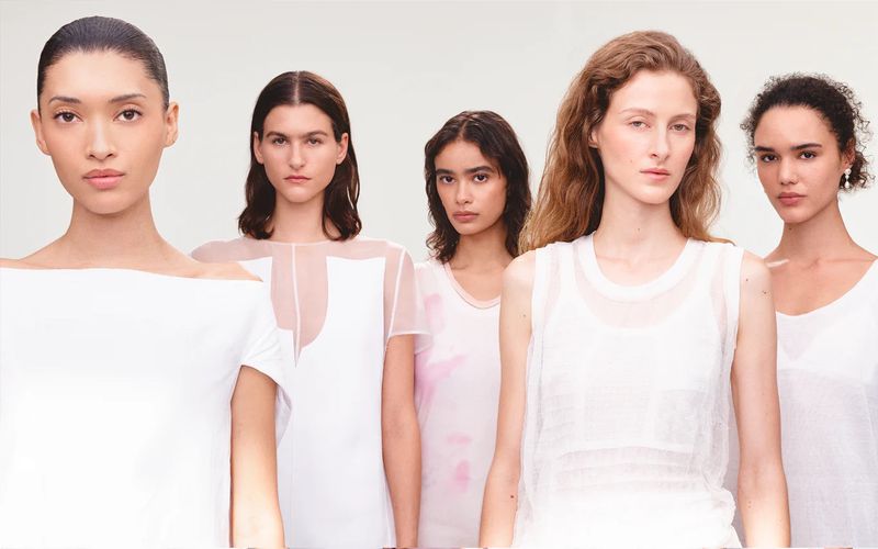 Five women pose in white tops against a bright background. The models face forward with natural makeup and a minimalist fashion aesthetic.