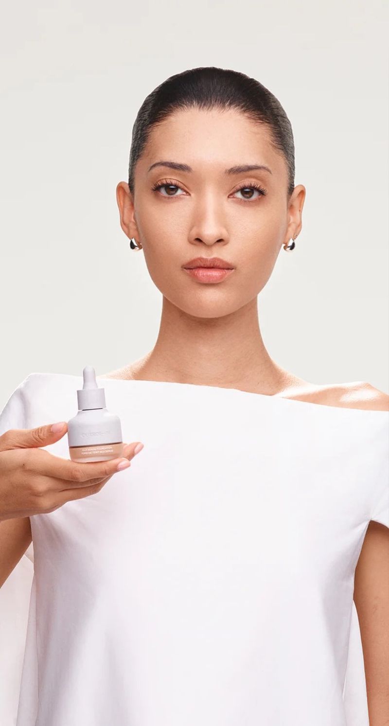 Adult female poses in a white top while holding a foundation bottle in an indoor photo studio setting with soft lighting.