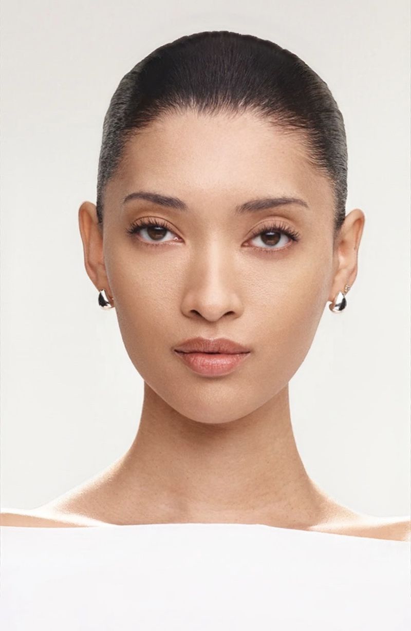 Close-up studio shot of a woman with dark hair, wearing earrings, against a clean white background. Portrait photograph for beauty or fashion.