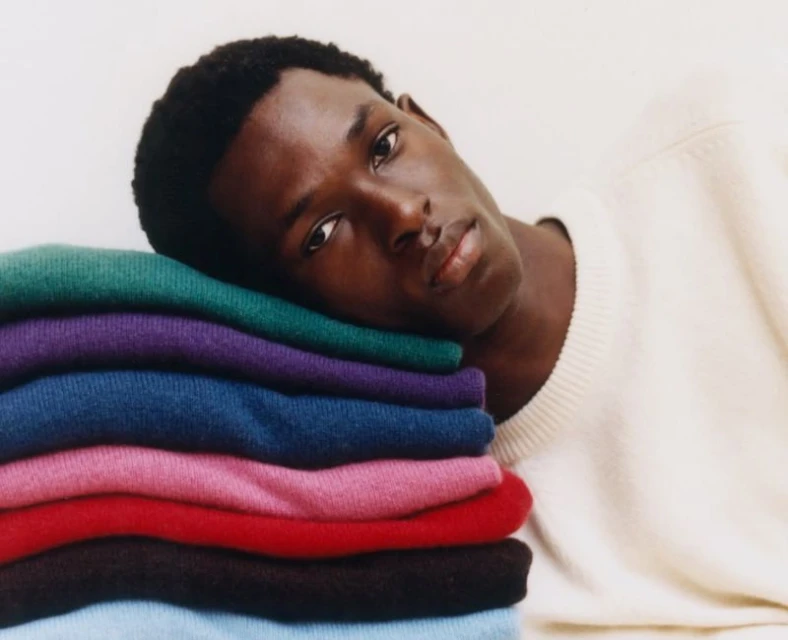 Portrait of a Black male model leaning on a stack of colorful sweaters, wearing a cream sweater, showcasing fashion and style.