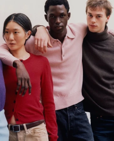 Three people standing together in a studio: a red cardigan on a woman, a pink polo shirt on a man, and a brown sweater on another man.
