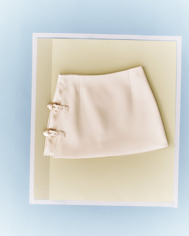 Ivory mini skirt with buckle details is framed by a layered white and beige paper border against a pale blue background in a studio shot.