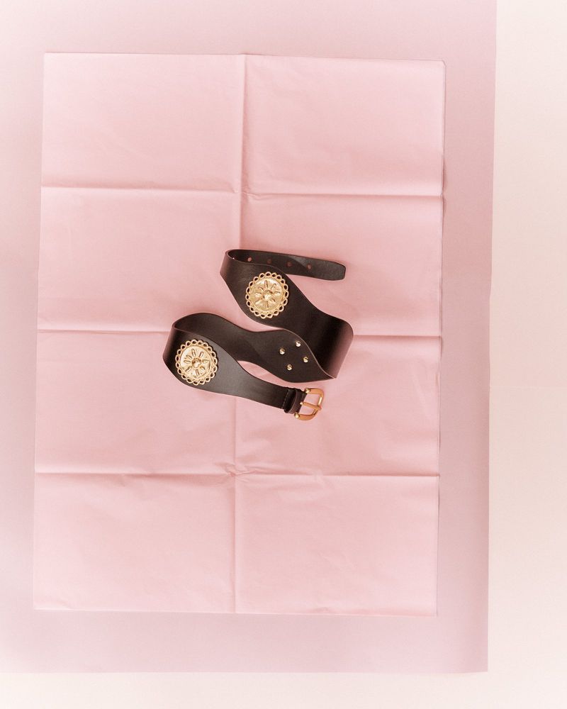 Flat lay studio shot of a black leather belt with gold accents arranged on pink paper; fashion accessory, studio lighting.