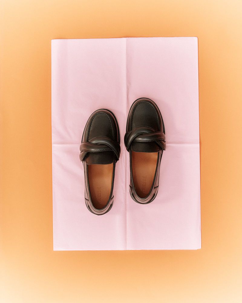 Overhead studio shot of a pair of black leather twisted knot loafers on a folded pink tissue paper background against a peach backdrop.