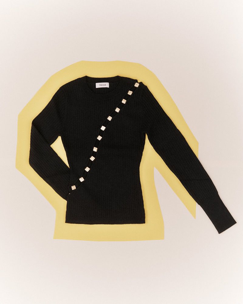 Studio shot of a long-sleeve, black, ribbed, knit shirt with decorative buttons, lying on a yellow backdrop over a white surface.