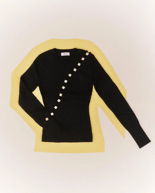 Studio shot of a long-sleeve, black, ribbed, knit shirt with decorative buttons, lying on a yellow backdrop over a white surface.